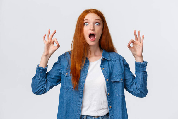 Amused, excited and thrilled good-looking redhead female in denim shirt, open mouth, drop jaw speechless staring with popped eyes camera, showing okay gesture in approval, white background - 写真・画像