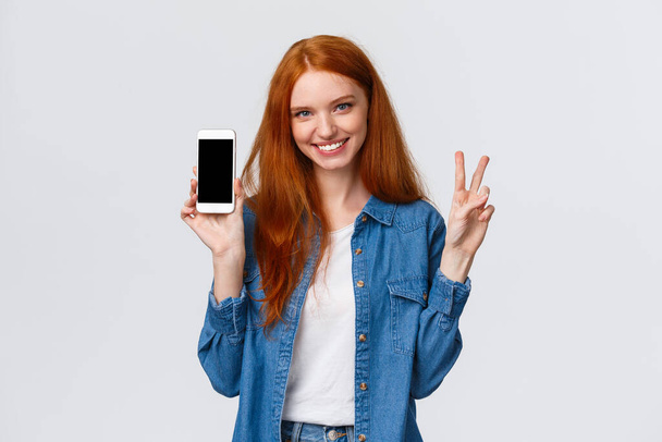 Χαριτωμένη και χαρούμενη σαγηνευτική κοκκινομάλλα γυναίκα με denim πουκάμισο, που δείχνει την ειρήνη χειρονομία χαμογελώντας και κρατώντας smartphone, εμφάνιση app, ιστοσελίδα της εταιρείας ή κατάστημα στο διαδίκτυο για ψώνια στην οθόνη του κινητού, λευκό φόντο - Φωτογραφία, εικόνα