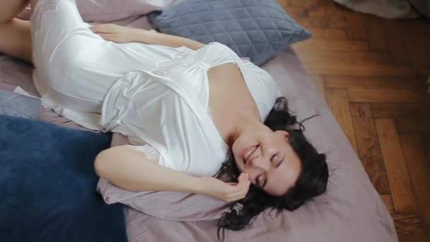 Menina jovem europeia bonita a noiva em roupa interior branca e peignoir encontra-se na cama na pose sexual e gosta da manhã, sorrindo do prazer com olhos fechados. Sessão de fotos em estúdio de noiva bonito
 - Filmagem, Vídeo