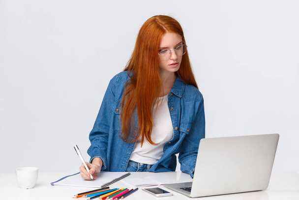 Inspiraatio, työ ja taidekonsepti. Keskittynyt kiireinen söpö punapää nainen lasit, kirjoittaa muistiinpanoja kannettavasta tietokoneesta, käytä hyödyllistä verkkosivustoa, verkkokurssi oppia piirustus, valkoinen tausta - Valokuva, kuva