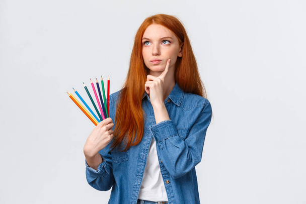 Gondolkodó és koncentrált, komoly kinézetű, eltökélt vörös hajú diáklány, tinédzser akar rajzolni valami érdekeset, színes ceruzákat tartva felnéz gondolkodás, elmélkedő ötletek - Fotó, kép
