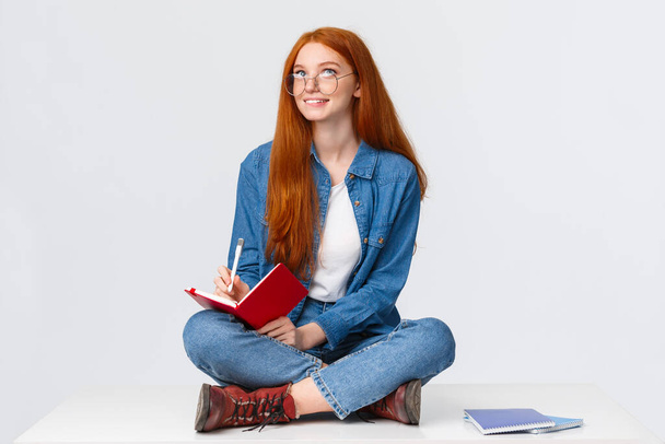 Dreamy and creative cute diligent female student with red hair, in glasses, sitting on floor with legs crossed surrounded by notebooks writing something, creating poem, look up thoughtful - Zdjęcie, obraz