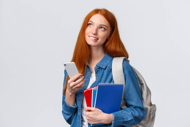 Dreamy and cute good-looking redhead imaging girl, biting lip from desire or temptation, looking thoughtful up, smiling holding backpack, notebooks and smartphone, chatting friends - Photo, image