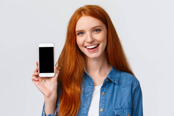 Close-up portret moderne millennial schattige roodharige vrouw met blauwe ogen, advies krijgen cool nieuwe app, tonen smartphone display en glimlachen, reclame spel of foto filter applicatie - Foto, afbeelding