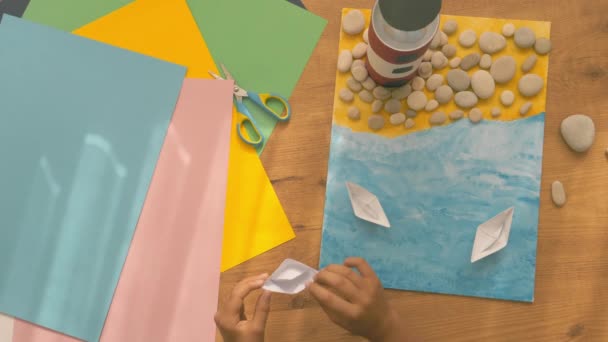 Kid hands make a lighthouse for school project at home. Top down view to wooden table with colored paper and other supplies - Footage, Video