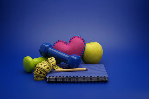 Fitness en gewichtsverlies concept met halters, meetlint, hart, groene appel en nieuwe notebook op blauwe achtergrond - Foto, afbeelding