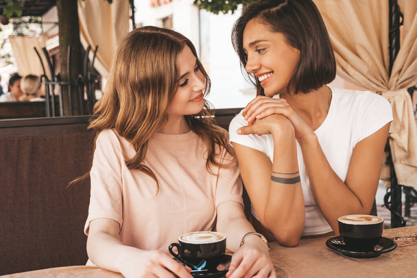 Two young beautiful smiling hipster girls in trendy summer casual clothes.Carefree women chatting in veranda terrace cafe and drinking coffee.Positive models having fun and communicating - Φωτογραφία, εικόνα