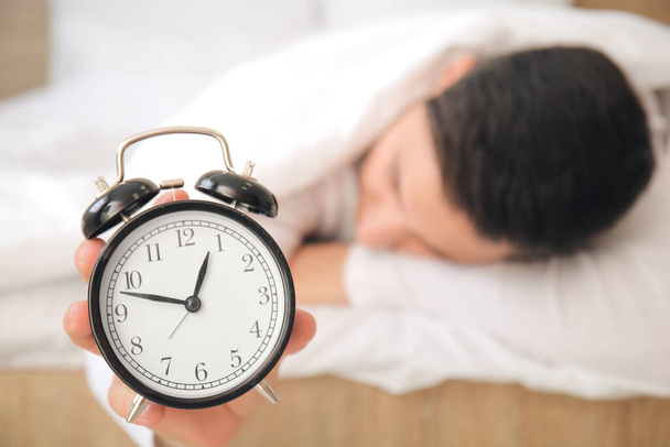 Young man with alarm clock falling asleep in bedroom. Concept of sleep deprivation - Φωτογραφία, εικόνα