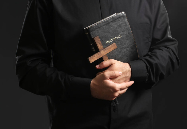 Young priest with Bible and cross on dark background - Photo, Image
