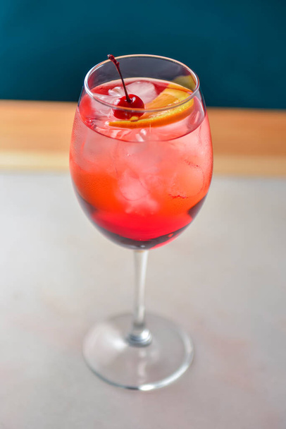 Citrus fruit cocktail in nice red color in front of a black background. Served in a wine glass on a wooden table. Refreshing alcohol summer drink with ice and cherry on a wooden table - Φωτογραφία, εικόνα