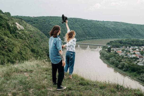 Joven pareja viajera de moda divirtiéndose en la cima de la colina disfrutando de la puesta de sol sobre el valle cerca del río, viajes y senderismo Concepto de viaje
 - Foto, Imagen