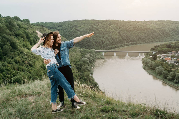 Joven pareja viajera de moda divirtiéndose en la cima de la colina disfrutando de la puesta de sol sobre el valle cerca del río, viajes y senderismo Concepto de viaje
 - Foto, Imagen