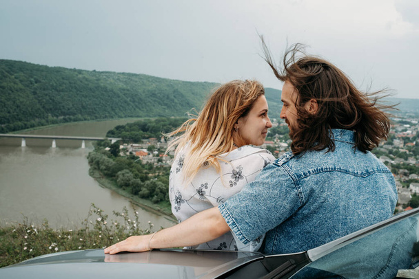 Jeune couple de voyage à la mode s'amusant près de la voiture au sommet de la colline Voyage et Road Trip Concept - Photo, image