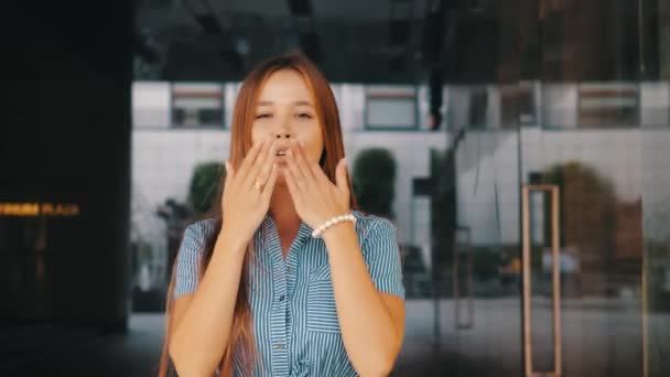 Front front view of charming cute smiling long-haired girl in striped dress sending air kisses to camera against glass reflective wall of restaurant. Positive happy young woman conveys kisses of love. - Footage, Video