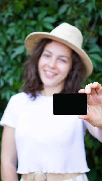 Frau hält leere Kreditkarte für Attrappe. Verschwommen lächelndes Mädchen mit Sommermütze und weißem T-Shirt vor ihrer schwarzen EC-Karte, in die Kamera blickend, auf grünem Hintergrund im Freien. Vertikal.  - Foto, Bild