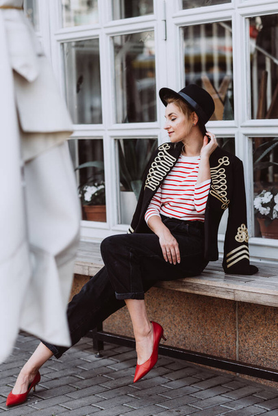 Stijlvolle jonge vrouw blond in een hussar jas, zwarte hoed, jeans, bananen en rode schoenen in de stad bij het raam van het restaurant. Moderne mode. Zachte selectieve focus. - Foto, afbeelding