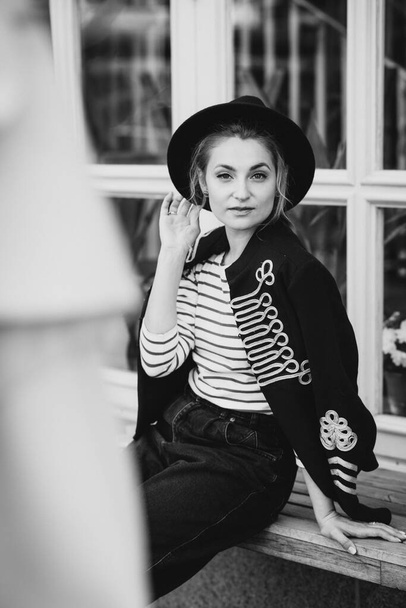 スタイリッシュな若い女性のブロンドは、レストランの窓の近くの街の黒い帽子のハッサージャケット。モダンなファッション。柔らかい選択的フォーカス. - 写真・画像
