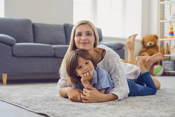 Loving mother hugging her son while lying on carpet in living room. Parent and child embracing at home. Family bonding - Valokuva, kuva