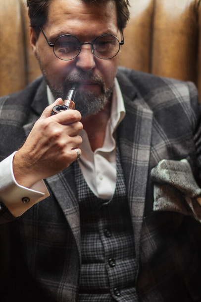 Reflective well-dressed mature bearded man in glasses smoking tobacco pipe - Foto, immagini