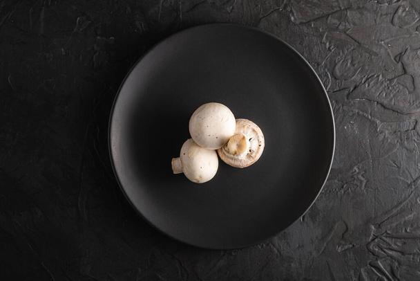 Three champignon mushrooms healthy food on black plate on dark black textured background, top view - Photo, Image