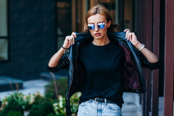 Ragazza bionda che indossa t-shirt nera, occhiali e giacca di pelle in posa contro strada, stile di abbigliamento urbano. Foto di strada - Foto, immagini