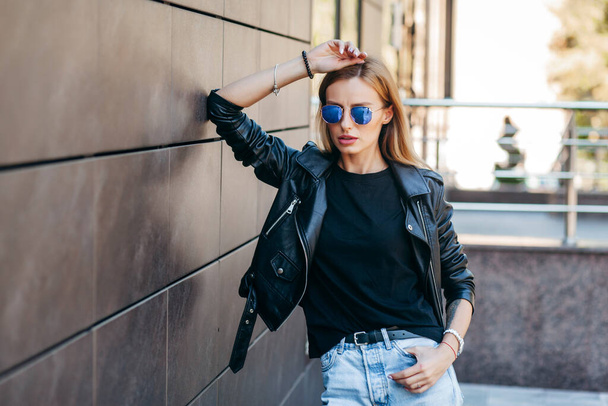 Blonde girl wearing black t-shirt, glasses and leather jacket posing against street , urban clothing style. Street photography - Photo, Image