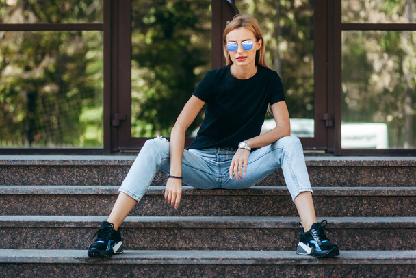 Élégante fille blonde portant un t-shirt noir et des lunettes posant contre la rue, style vestimentaire urbain. Photographie de rue - Photo, image