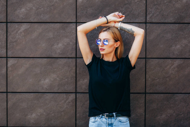 stijlvol blond meisje met zwart t-shirt en bril poserend tegen de straat, stedelijke kledingstijl. Straatfotografie - Foto, afbeelding