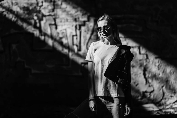 Girl wearing t-shirt, glasses and leather jacket posing against street , urban clothing style. Street photography - Photo, Image