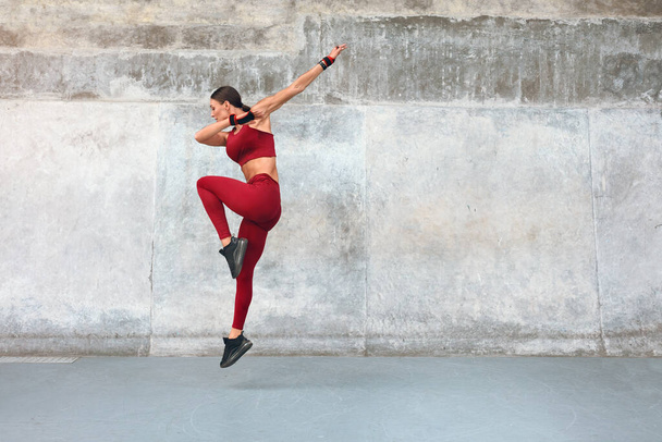 Fitness. Jumping Woman Doing Workout At Outdoor Stadium. Side View Of Fit Girl With Strong Muscular Body In Fashion Sporty Outfit Doing Exercise Against Concrete Wall. Dynamic Movement. - Foto, imagen