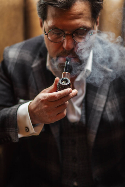 Reflective well-dressed mature bearded man in glasses smoking tobacco pipe - Fotó, kép