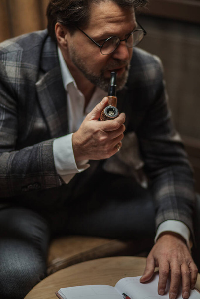 Fifty years old businessman making notes on the paper, smoking a tobacco pipe - Фото, изображение