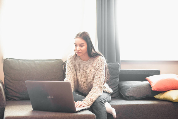 Young woman with modern laptop sitting on sofa at home. Happy girl browsing through the internet during free time..  - Zdjęcie, obraz