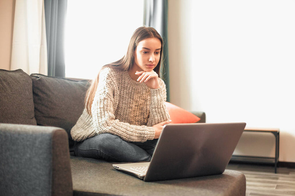 Young woman with modern laptop sitting on sofa at home. Happy girl browsing through the internet during free time..  - Φωτογραφία, εικόνα