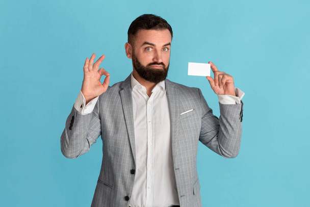 Confident corporate employee holding empty business card and showing ok gesture on blue background, space for design - Fotografie, Obrázek