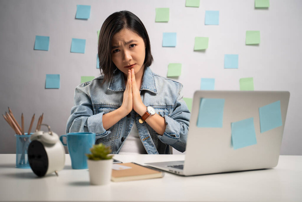 祈りの中で手をつないでいるアジアの女性は、カメラを見て、自宅のオフィスでノートパソコンに取り組んでいます。WFH 。家から仕事だ。予防コロナウイルスCOVID-19の概念. - 写真・画像