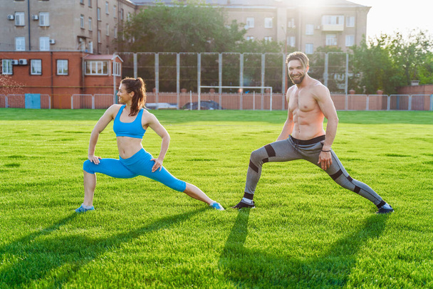 Jeune couple attrayant homme et femme de 25 ans font des exercices sur un matin d'été ensoleillé sur le stade de sports d'herbe. Joyeux et souriant faisant des pentes, étirant les muscles, levant les jambes. - Photo, image