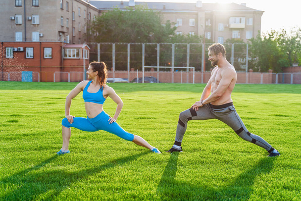 Attractive young couple man and woman 25 years old are doing exercises on a sunny summer morning on grass sports stadium. Joyfully and with a smile making slopes, stretching muscles, raising legs. - 写真・画像