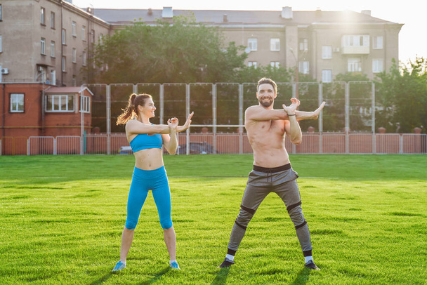 Attractive young couple man and woman 25 years old are doing exercises on a sunny summer morning on grass sports stadium. Joyfully and with a smile making slopes, stretching muscles, raising legs. - Fotoğraf, Görsel