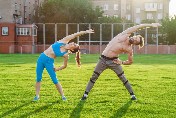 魅力的な若いカップルの男性と女性25歳は、芝生のスポーツ競技場で晴れた夏の朝に演習を行っています。喜びと笑顔で斜面を作り、筋肉を伸ばし、足を上げる. - 写真・画像