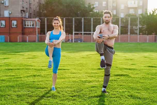 Привлекательная молодая пара мужчина и женщина 25 лет делают упражнения солнечным летним утром на травяном спортивном стадионе. Радостно и с улыбкой делая склоны, растягивая мышцы, поднимая ноги
. - Фото, изображение