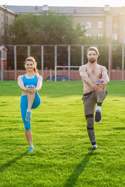 Atraente jovem casal homem e mulher de 25 anos estão fazendo exercícios em uma manhã de verão ensolarada no estádio de esportes de grama. Alegremente e com um sorriso fazendo encostas, alongando músculos, levantando pernas
. - Foto, Imagem