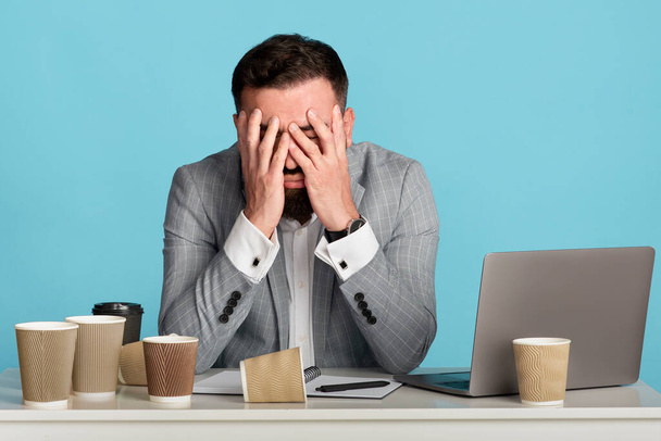 Empresário exausto tentando não dormir no local de trabalho com xícaras de café vazias e laptop no fundo azul
 - Foto, Imagem