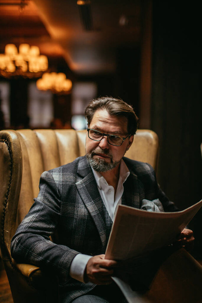 Mid-aged well-dressed man sitting in arm-chair in living room reads newspaper - Φωτογραφία, εικόνα