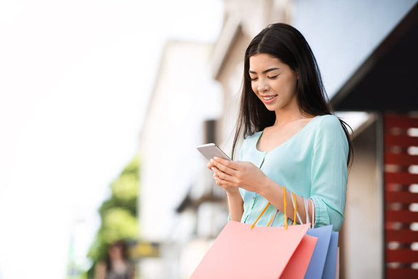 Moda satışları ve anlaşmaları. Alışveriş çantalı Asyalı kız dışarıda akıllı telefon kullanıyor. - Fotoğraf, Görsel