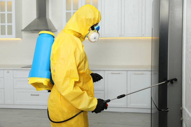 Pest control worker in protective suit spraying insecticide near refrigerator indoors - Foto, immagini