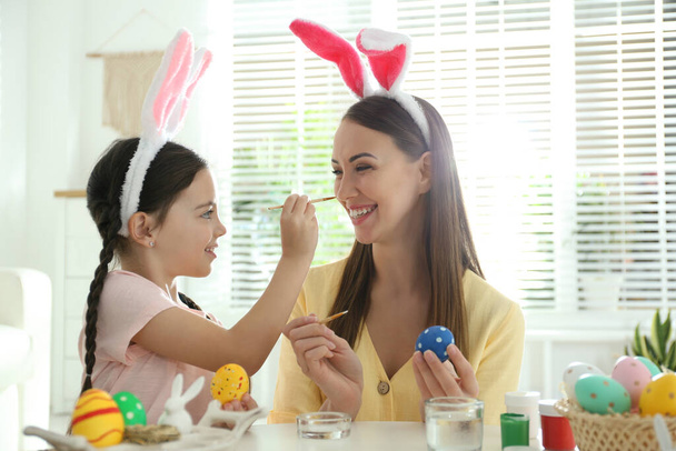 Happy mother and daughter with bunny ears headbands having fun while painting Easter eggs at home - Foto, afbeelding