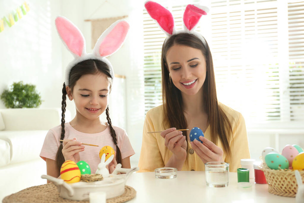Happy mother and daughter with bunny ears headbands painting Easter eggs at home - Фото, зображення