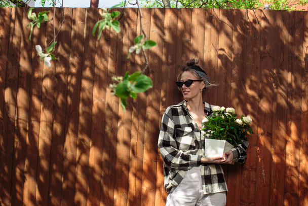 Молодая красивая девушка в стильной одежде прислоняется к деревянному забору и держит цветочный горшок с порезанной розой
. - Фото, изображение