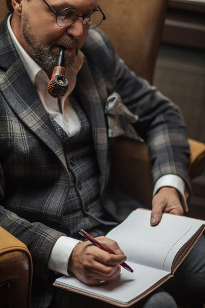 Mature businessman smoking tobacco pipe and making notes, working at his office. - Foto, Imagem